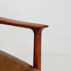 May include: Close-up of the armrest of a brown leather chair with a wooden frame. The armrest is made of solid wood and has a smooth, curved design.