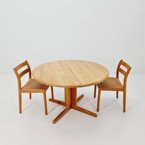 Könnte beinhalten: Ein runder Esstisch aus hellem Holz mit einem Säulenfuß, flankiert von zwei passenden Stühlen. Die Stühle haben gewebte Sitze und Sprossenrücken. Der Tisch hat einen Durchmesser von etwa 122 cm.