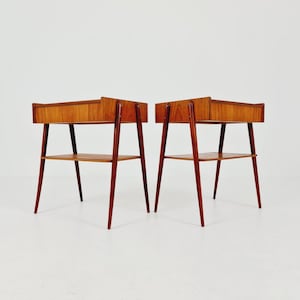 Op de afbeelding: Een paar houten bijzettafels met een rechthoekig blad met opstaande rand, een enkele lade en een onderste plank. Elke tafel wordt ondersteund door vier uitlopende, taps toelopende poten en is afgewerkt met een natuurlijke houtnerf.