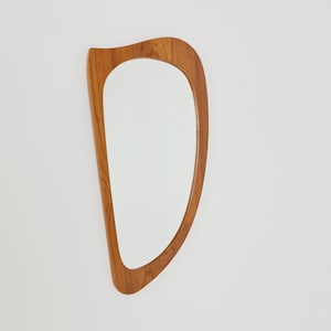 Può includere: Uno specchio da parete con una cornice asimmetrica dalla forma organica, realizzata in legno marrone chiaro con venature visibili. Il vetro dello specchio è racchiuso nel bordo in legno curvo, creando una silhouette fluida e non geometrica.