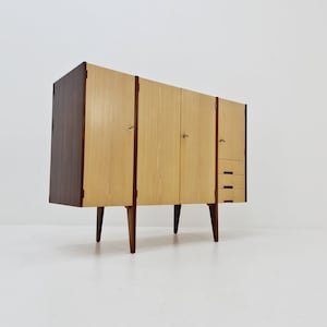 Op de afbeelding: Een modern dressoir uit het midden van de eeuw met drie deuren en drie laden. De kast is gemaakt van hout met een lichte houtkleur en donkerhouten afwerking. Het heeft vier poten en staat op een wit oppervlak.