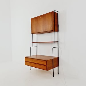 Może przedstawiać: Montowany na ścianie regał w stylu mid-century modern. Posiada szafkę, półkę i szuflady z jasnego drewna. Konstrukcja wsparta jest na czarnych metalowych prętach. Szafka ma jedne drzwi, a szuflady dwie szuflady.