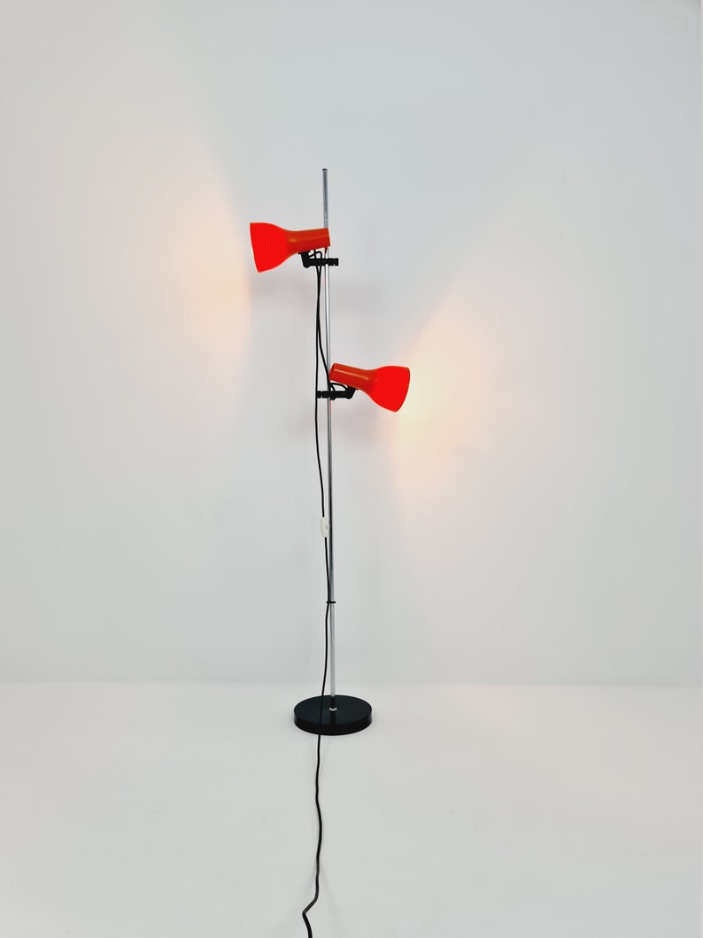 Puede incluir: Una l&aacute;mpara de pie de cromo con dos pantallas rojas ajustables. La l&aacute;mpara tiene una base negra y un cable negro largo.