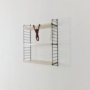 May include: A three-tiered wall-mounted shelf with a black metal frame and light wood shelves. A small brown monkey figurine hangs from the top shelf. The shelves are attached to the wall with black metal supports.