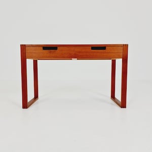 Op de afbeelding: Een rechthoekig houten bureau met een warme bruine houtnerfafwerking. Het bureau bevat twee naast elkaar geplaatste lades met zwarte verzonken horizontale handgrepen. Het wordt ondersteund door vier rechte poten verbonden door twee U-vormige zijframes.