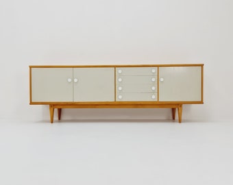 May include: A mid-century modern sideboard with a light wood frame and white doors and drawers. The sideboard has a simple design with rounded edges and small, round knobs.