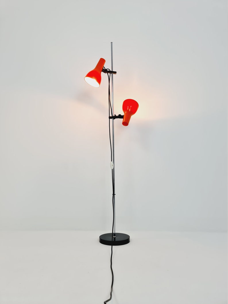 Puede incluir: Una l&aacute;mpara de pie cromada con dos pantallas de l&aacute;mpara naranja ajustables. La l&aacute;mpara tiene una base negra y un cable negro largo.