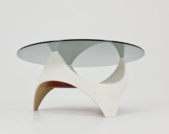 May include: A round coffee table featuring a circular, smoked gray glass top supported by a sculptural, off-white base. The base consists of three curved, interconnected legs that create a hollow, open center with a warm wood-toned interior.
