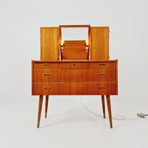 May include: A wooden vanity dresser featuring three horizontal drawers with recessed handles, supported by four tapered legs with brass-toned feet. The top section includes a central mirror flanked by two wooden panels and two integrated light fixtures.