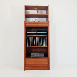 Könnte beinhalten: Ein hoher Schallplattenschrank aus Holz mit mehreren Regalen und einer Schublade. Der Schrank hat eine warme braune Farbe und verfügt über einen eingebauten Plattenspieler. Eine kleine Holzfigur eines Affen hängt am obersten Regal.