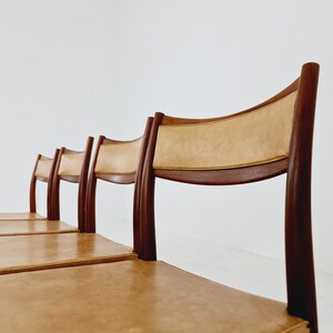 May include: Four brown wooden chairs with tan leather seats. The chairs are lined up in a row.