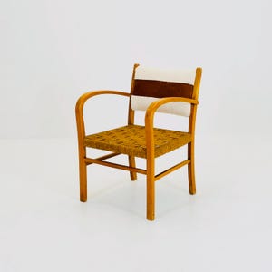 May include: A vintage wooden armchair with a woven seat and back. The chair features curved armrests and a backrest with a brown leather and cream-colored fabric panel. The chair is made of light-colored wood.