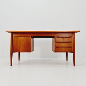 May include: A mid-century modern wooden desk with a warm, reddish-brown finish. The desk features a rectangular top, a cabinet with a door on the left, and three drawers on the right. The desk has tapered legs and a keyhole on the drawer.