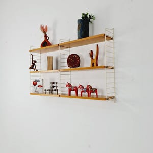 May include: A white metal shelving unit with wooden shelves. The shelves are filled with various decorative items, including a vase with dried flowers, a clock, a wooden cat figurine, and three red wooden horses. The shelves are mounted on a white wall.