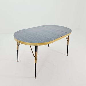 May include: An oval mid-century modern table with a grey wood grain top and brass-coloured trim. The table has four black and brass legs. A stylish piece of furniture for a living or dining area.