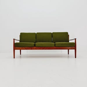 Puede incluir: Un sofá de tres plazas de estilo moderno de mediados de siglo, con estructura y patas de madera. Los cojines son de un verde oliva oscuro, que contrasta con el marrón cálido de la madera. El sofá tiene un diseño minimalista.