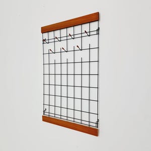 May include: A wall-mounted organizer with a grid of black metal wires and wooden trim. The organizer has several small hooks with red tips. The wooden trim is a warm brown color.