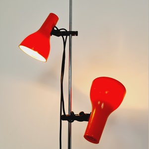 Puede incluir: Una l&aacute;mpara de pie roja con dos cabezales ajustables. La l&aacute;mpara tiene un poste cromado y cables negros.