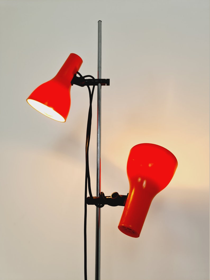 Puede incluir: Una l&aacute;mpara de pie roja con dos cabezales ajustables. La l&aacute;mpara tiene un poste cromado y cables negros.