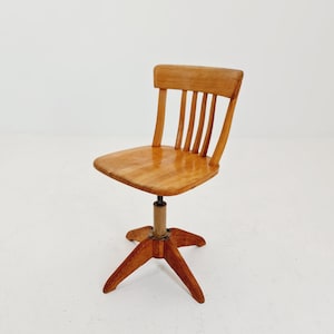 Könnte beinhalten: Ein Vintage-Schreibtischstuhl aus hellem Holz. Der Stuhl hat eine Rückenlehne mit Sprossen, einen quadratischen Sitz und ein Fünf-Punkt-Gestell. Der Stuhl ist drehbar und höhenverstellbar.