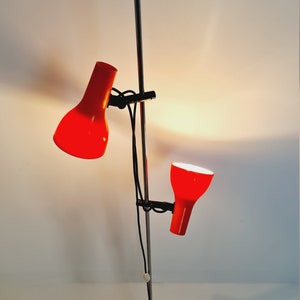 Puede incluir: Una l&aacute;mpara de pie de cromo con dos pantallas rojas ajustables. La l&aacute;mpara tiene una base negra y un poste largo y delgado de cromo.