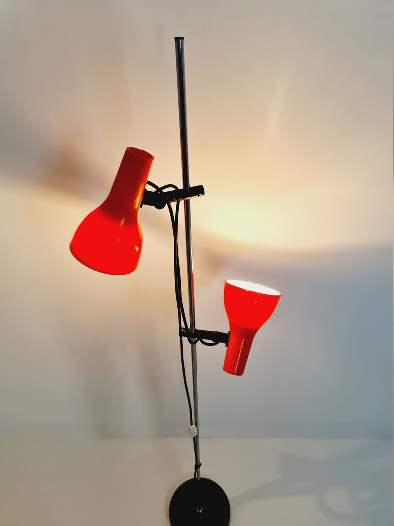 Puede incluir: Una l&aacute;mpara de pie de cromo con dos pantallas rojas ajustables. La l&aacute;mpara tiene una base negra y un poste largo y delgado de cromo.