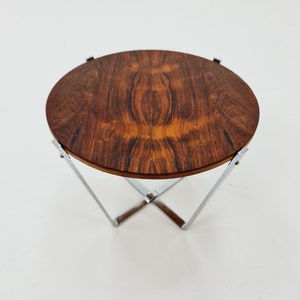 Könnte beinhalten: Ein runder Holztisch mit einem verchromten Metallgestell. Die Tischplatte ist aus dunklem Holz mit natürlicher Maserung. Das Gestell ist ein einfaches X-förmiges Design.