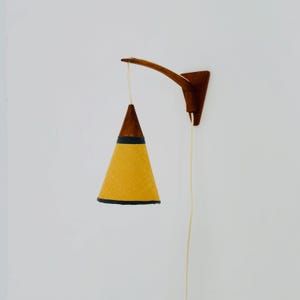 Könnte beinhalten: Vintage Wandleuchte mit Holzarm und Halterung. Die Lampe hat einen gelben Stoffschirm mit schwarzem Rand und eine dunkelbraune Holzspitze. Ein cremefarbenes Kabel hängt herunter. Das Design ist modern.
