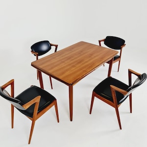 Puede incluir: Una mesa de comedor rectangular de madera con un acabado de veta suave y cuatro sillas a juego. Cada silla presenta una estructura de madera con reposabrazos angulados y un asiento y respaldo tapizados en color negro.