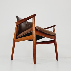 May include: A brown leather armchair with a wooden frame. The chair has a simple design with a slightly curved back and armrests. The chair is sitting on a white floor.