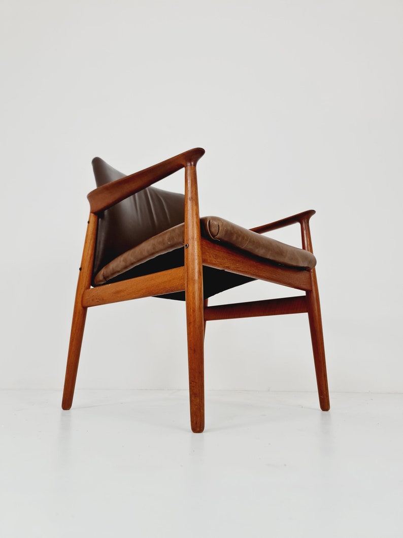 May include: A brown leather armchair with a wooden frame. The chair has a simple design with a slightly curved back and armrests. The chair is sitting on a white floor.