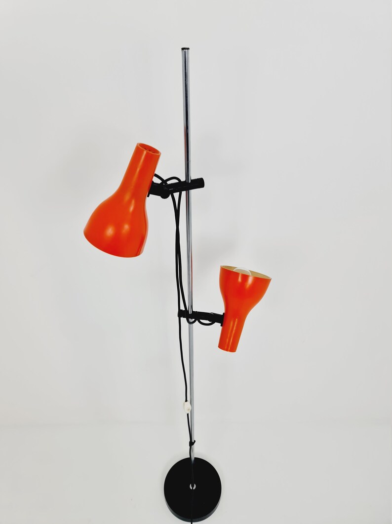 Puede incluir: Una l&aacute;mpara de pie de cromo con dos pantallas de l&aacute;mpara naranja ajustables. La l&aacute;mpara tiene una base negra y un poste de cromo largo y delgado.