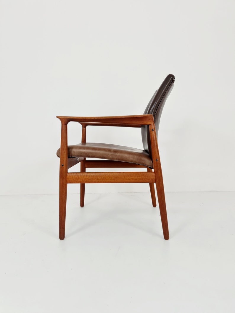 May include: A brown leather chair with a wooden frame. The chair has a high back and arms. The chair is sitting on a white surface.