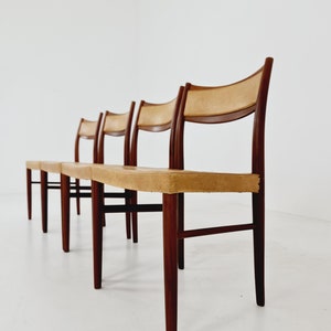 May include: Four wooden chairs with tan leather seats. The chairs are arranged in a row, with the front chair in focus. The chairs have a simple, mid-century modern design.