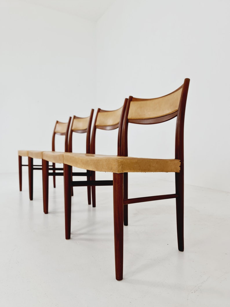 May include: Four wooden chairs with tan leather seats. The chairs are arranged in a row, with the front chair in focus. The chairs have a simple, mid-century modern design.