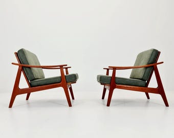 May include: Two mid-century modern armchairs. The chairs have a reddish-brown wooden frame and dark green cushions. The armrests are curved, and the chairs are set against a white background.