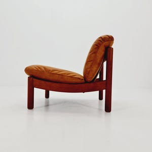 May include: A low-profile lounge chair featuring a dark brown wooden frame with four cylindrical legs and a curved backrest. The seat and backrest are fitted with plush, tan-colored leather cushions that provide a soft, rounded appearance.