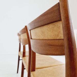May include: Four wooden chairs with light brown upholstery and a dark brown wood frame. The chairs are arranged in a row, with the back of the chair in the foreground.
