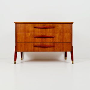 Puede incluir: Una cómoda de tres cajones de mediados de siglo con un acabado cálido de vetas de madera. Tiene tiradores y patas rojos con puntas color latón. El diseño es sencillo y elegante, adecuado para un dormitorio o sala de estar. Aproximadamente 80 cm de ancho.