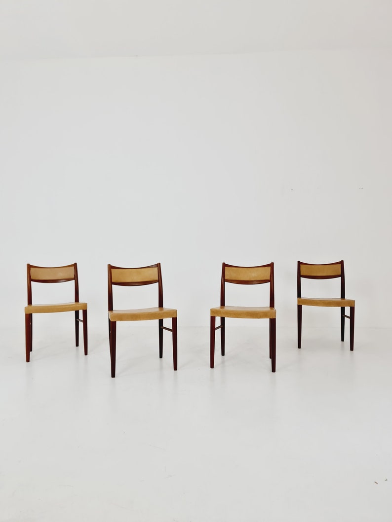 May include: Four chairs with a light brown leather seat and dark wood legs. The chairs are arranged in a row on a white floor.