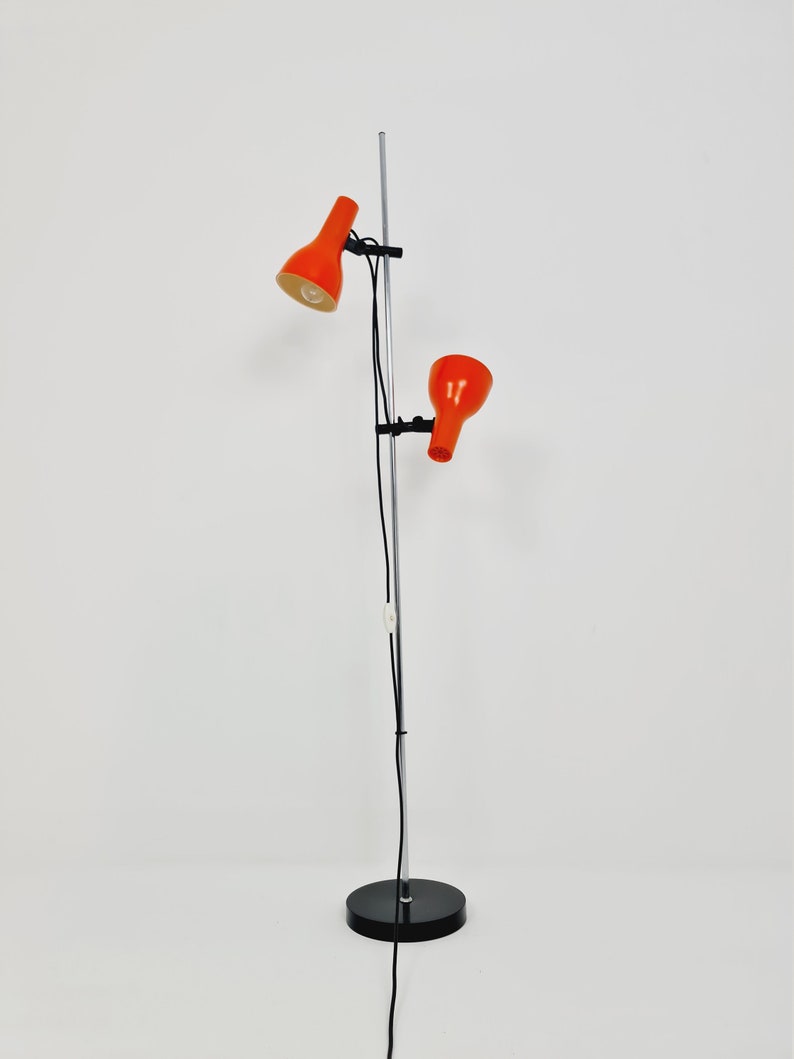 Puede incluir: Una l&aacute;mpara de pie de cromo con dos pantallas ajustables de color naranja. La l&aacute;mpara tiene una base negra y un cable largo.