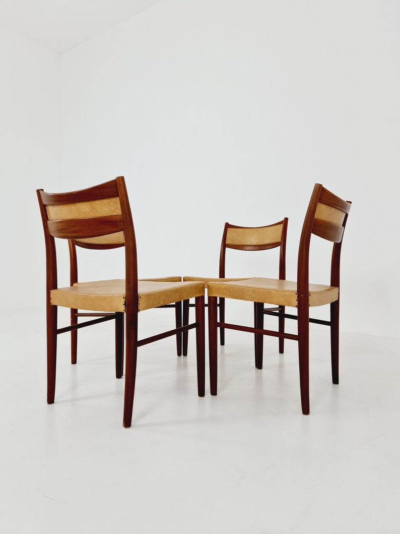 May include: Four wooden chairs with tan leather seats. The chairs are arranged in a square formation, with the backs of the chairs facing the camera.