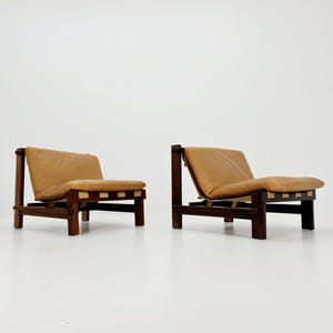 May include: Two mid-century modern armchairs. Each chair has a dark wood frame with a light brown leather seat and back. The seats are supported by light-colored straps. The chairs are set against a white background.