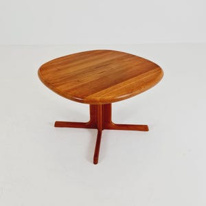 Op de afbeelding: Een middelbruine houten bijzettafel met een vierkant blad met afgeronde hoeken en randen. De tafel wordt ondersteund door een centrale kolomvoet die zich vertakt in vier afzonderlijke, slanke poten die vanuit het middelpunt naar buiten lopen.