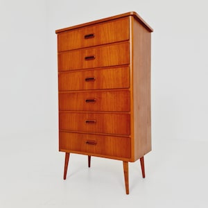 May include: A tall wooden chest of drawers featuring six horizontal drawers with integrated oval-shaped recessed handles. The piece is constructed from a warm-toned wood with a visible grain pattern and is supported by four tapered wooden legs.