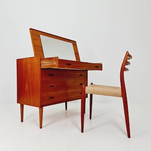 Op de afbeelding: Een houten dressoir in mid-century modern stijl met een ingebouwde spiegel en drie lades. Een bijpassende houten stoel met een geweven zitting staat naast het dressoir. Het meubilair is gemaakt van warm getint hout.