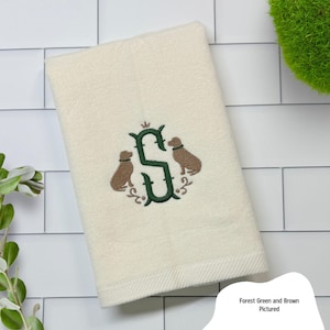 May include: Cream-colored hand towel with an embroidered design. The design features a forest green letter "S" with a crown, flanked by two brown dog silhouettes, and floral accents. The towel is folded and has a textured border.