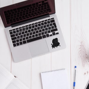 May include: A silver laptop computer with a black keyboard and a black silhouette of a poodle on the top right corner of the laptop. The laptop is on a white wooden surface.
