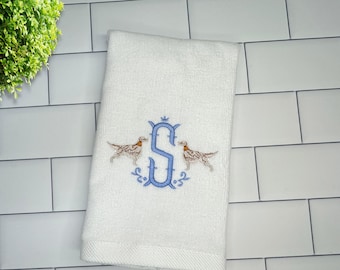 May include: White hand towel with an embroidered design. The design features a blue letter "S" with two brown and white dogs on either side. The towel is folded and placed on a white tiled surface.