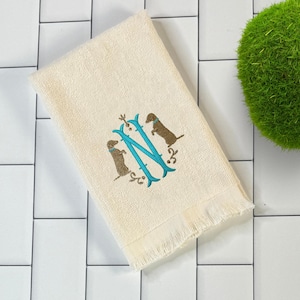 May include: Cream-colored hand towel with a fringed edge. The towel features an embroidered teal letter "N" with two brown dachshunds and decorative floral accents. The towel is placed on a white tiled surface.